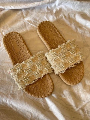 Beige sandaler med pärlor och fransar - Säljer ett par beige sandaler med flätad sula och bred rem i tyg dekorerad med pärlor och fransar. Sandalerna har en bohemisk vibe och är perfekta för sommaren. Materialet är en mix av textil och syntet, och designen är öppen med slip-in modell.