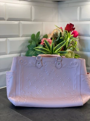 Ljusrosa Louis Vuitton handväska - Säljer en ljusrosa handväska från Louis Vuitton med präglat monogrammönster och silverfärgade detaljer. Väskan har en rektangulär form, dubbla handtag och är tillverkad i glansigt läder. Perfekt för dig som vill ha en lyxig och trendig accessoar.