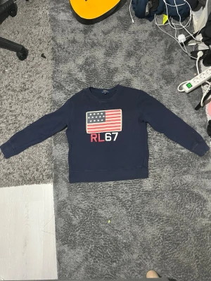 Mörkblå sweatshirt Polo Ralph Lauren - Cool mörkblå sweatshirt från Polo Ralph Lauren med amerikansk flagga och RL67 tryck på bröstet. Klassisk rund hals och långa ärmar, perfekt för en avslappnad och stilren look.