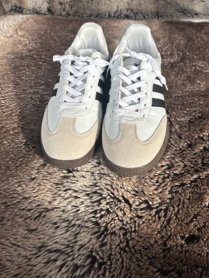 Adidas Samba vita och svarta sneakers - Klassiska Adidas Samba sneakers i vitt läder med svarta ränder och beige mockadetaljer framtill. Yttersulan är brun och har ett mönster för bra grepp. Skorna har vita skosnören och låg profil som ger en clean och tidlös look. Använda ett få tal gånger då jag köpte dom för 2 år sen men det var inte riktigt min stil.