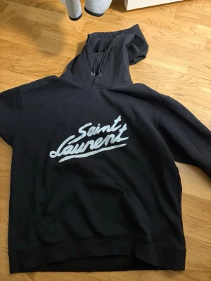 Svart Saint Laurent hoodie - Säljer en svart hoodie från Saint Laurent med stor vit logga på bröstet. Hoodien har huva med dragsko och en klassisk magficka framtill. Perfekt för dig som gillar streetwear och vill ha något stilrent men ändå lyxigt.