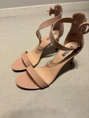 Nude lackpumps med ankelrem - Snygga pumps i nude färg med smal rem över tårna och justerbar ankelrem med spänne. Skorna har smal klack och är tillverkade i glansigt lackmaterial som ger en elegant look. Perfekta för dig som vill ha en stilren och trendig sko med öppen tå.