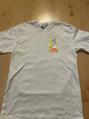 Vit t-shirt från Casablanca - Säljer en vit t-shirt från Casablanca med tryck i gult, blått och rött på bröstet där det står 'Casa Blanca'. T-shirten har rund hals och korta ärmar, tillverkad i mjuk bomull. Perfekt för dig som gillar stilrena plagg med en twist.