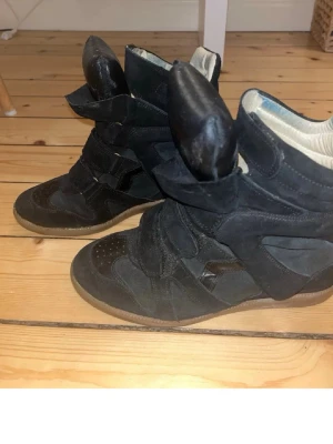 Isabel Marant skor - Säljer ett par svarta höga sneakers från Isabel Marant med unik design och kraftig vadderad plös. Skorna har en mix av mocka och läder, kardborreband och vit vadderad insida. Perfekta för dig som vill sticka ut med din stil.