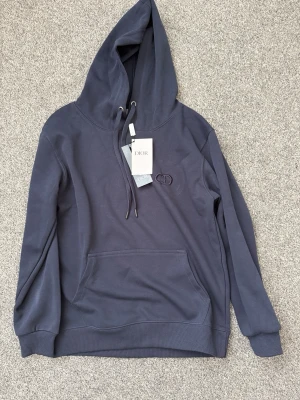 Mörkblå hoodie från Dlor - Snygg mörkblå hoodie från Dlor med klassisk huva och dragsko. Broderad CD-logga på bröstet och stor magficka framtill. Tillverkad i mjuk bomull som känns riktigt skön mot huden. Perfekt för dig som vill ha en stilren och exklusiv look.