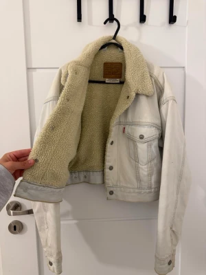 Vit sherpa jeansjacka från Levi's - Snygg vit jeansjacka från Levi's med mjukt och varmt sherpa-foder. Klassisk modell med två bröstfickor, knappar framtill och detaljer i silver. Perfekt för dig som vill ha en cool och tidlös look med extra komfort.