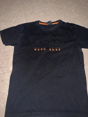 Svart Hugo Boss t-shirt med logga - Snygg svart t-shirt från Hugo Boss med broderad BOSS-logga och texten HUGO BOSS i orange på bröstet. Klassisk rund halsringning med orange detalj på insidan av kragen. Perfekt för dig som gillar stilrena märkesplagg.