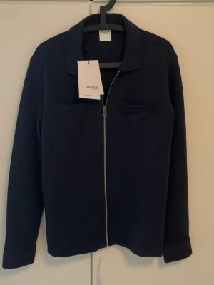 Mörkblå overshirt från Selected Homme - Stilren mörkblå overshirt från Selected Homme med dragkedja framtill och två bröstfickor. Klassisk krage och långa ärmar med manschett. Overshirten är tillverkad i ett mjukt tyg som ger en clean look och passar perfekt till sommarkväll