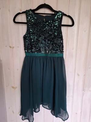 Mörkgrön finklänning stl 140 - Snygg mörkgrön klänning i stl 140, från H&M, med glittrig paljettöverdel och kjol i chiffong. Ärmlös modell med rund halsringning och markerad midja. Perfekt för kalas eller firande.