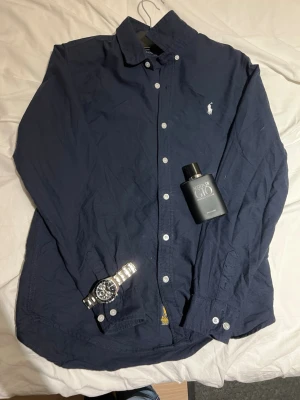 Mörkblå skjorta från Ralph Lauren - Klassisk mörkblå skjorta från Ralph Lauren med button-down krage och vita knappar. Skjortan har den ikoniska vita Polo-loggan broderad på bröstet och är tillverkad i ett mjukt bomullstyg. Perfekt till jeans för en clean och stilren look. Använd ca 2-3 gånger.