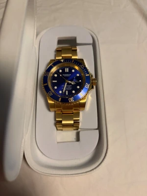 Blå och guldpläterad klocka från Kompass - Snygg klocka från Kompass med blå urtavla och guldfärgad länk. Klockan har tydliga indexmarkeringar, datumfönster och bezel i blått och guld. Klassisk design som sticker ut med sin färgkombination och metallarmband.