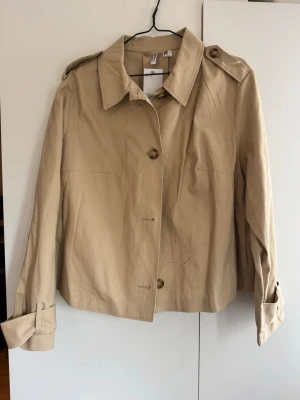 Kort beige trenchcoat från H&M - Stilren kort trenchcoat i beige från H&M med klassisk krage, axelklaffar och dekorativa manschetter. Jackan har stora knappar framtill och är tillverkad i ett lätt bomullsliknande material. Perfekt för dig som gillar minimalistisk och tidlös stil. Nypris 399 med tag. Helt ny 