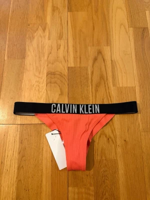 Rosa stringtrosa korallrosa från Calvin Klein Oanvänd XS - Snygg rosa stringtrosa från Calvin Klein med bred svart resår i midjan och tydlig logga framtill. Trosan har ribbad struktur och är tillverkad i ett mjukt, stretchigt material som sitter skönt på kroppen. Perfekt för dig som gillar sportig och trendig stil. Ordinarie pris 650 kr.