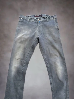  Jacob Cohën Jeans - Snygga blå slim fit jeans från Jacob Cohën med klassisk femficksdesign och diskret dekorerad bakficka. Jeansen har normal midja, bälteshällor och knappgylf. Materialet är mjukt denim som ger en bekväm känsla och en modern look. Perfekt skick förutom några vita pluppar som saknas på en av fickorna