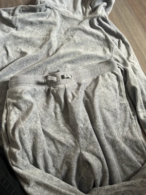 Grått loungewear-set Juicy Couture - Mysigt grått loungewear-set från Juicy Couture med hoodie och byxor. Setet har dragsko i midjan och diskreta strasstenar på både överdel och byxor. Tillverkat i mjukt material som passar perfekt för chill dagar hemma.