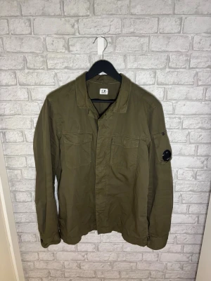 C.P. Company overshirt - C.P. Company overshirt med två bröstfickor och ikonisk linsdetalj på ärmen. Skjortkrage och knappstängning framtill. Tillverkad i lätt bomullsmaterial som passar perfekt för lager-på-lager. Hör av dig vid intresse 🙌