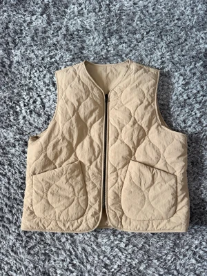 Beige quiltad dunväst med fickor - Stilren beige dunväst med quiltat mönster och två stora framfickor. Västen har dragkedja framtill och är ärmlös med rundad hals. Perfekt till sommar eller för lager på lager och ger en trendig look till din outfit.
