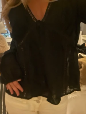 Svart blus med spetsdetaljer - Snygg svart blus med transparenta spetsdetaljer och knappar framtill. Blusen har v-ringning och långa ärmar med lätt puff. Materialet ser ut att vara tunn och luftig bomull eller viskos, perfekt för en trendig look. Super snygg både med och utan ett linne under, är köpt förra sommaren, storleken är L men tycker själv den sitter fint på mig som brukar jag ha xs-S i de flesta tröjorna beroende på märke så tror nog den passar S-L men kan sitta på olika sätt bara. Går att bära som en luftig kofta os