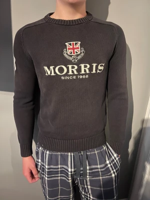 Svart stickad tröja från Morris - Snygg svart stickad tröja från Morris med Union Jack-emblem och texten 'Morris Since 1968' på bröstet. Stor vit 68-detalj på ena ärmen. Tröjan har rund halsringning och ribbade muddar. Perfekt för dig som gillar klassisk och sportig stil.