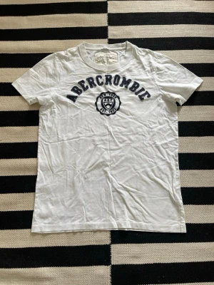 Vit Abercrombie t-shirt  - Klassisk vit t-shirt från Abercrombie & Fitch med mörkblå text och emblem på bröstet. T-shirten har rund hals, korta ärmar och är gjord i mjuk bomull. Perfekt till jeans eller shorts för en avslappnad look.