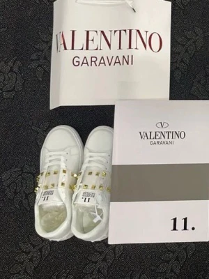 Valentino Rockstud vita sneakers - Snygga vita sneakers från Valentino Garavani, modell Rockstud Untitled. Skorna har coola guldfärgade nitar på ovandelen och klassisk låg siluett. Tillverkade i läder med vita snören och detaljer som sticker ut. Perfekta för dig som vill ha något extra.