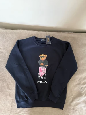  RLX Polo Bear sweatshirt  - Mörkblå sweatshirt från Polo Ralph Lauren med ikoniskt Polo Bear-tryck framtill. Björnen har solglasögon och rosa byxor, och RLX-logga syns under motivet. Tröjan har rund hals och långa ärmar. Perfekt för en avslappnad och cool stil. 