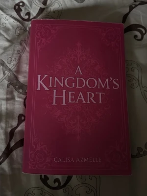 A Kingdom's Heart av Calisa Azmelle - En snyggt designad skönlitterär bok med vinröd mjukpärm och dekorativa mönster på omslaget. Titeln 'A Kingdom's Heart' är tryckt i stora, ljusa bokstäver. Perfekt för dig som gillar fantasy och romantik. Bokens rygg är knäckt, syns på andra och sista bilden.
