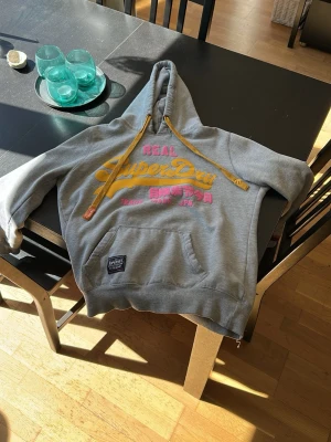 Grå Superdry hoodie med tryck - Säljer en grå hoodie från Superdry med stor gul och rosa logga på bröstet. Hoodien har gula dragsnören, känguruficka och mjukt fleecefoder. Perfekt för dig som gillar streetwear och vill ha något bekvämt och snyggt.