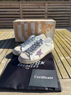 Vit sneakers med lila stjärna - Snygga vita sneakers från Golden Goose med en glittrig lila stjärna på sidan och leopardmönstrade skosnören. Skorna har beige mockadetaljer på tån och silverfärgad häl. Klassisk låg modell i läder som sticker ut med coola detaljer.