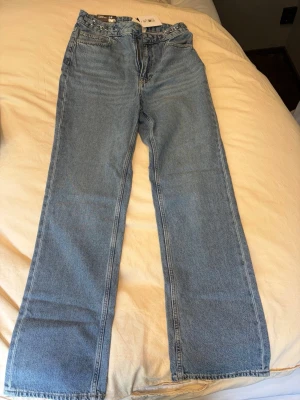 Blå raka jeans med kedjedetalj - Säljer ett par ljusblå raka jeans från divided med snygg kedjedetalj i midjan. Klassisk femficksmodell och hög midja. Jeansen är gjorda i bomullsdenim och har en avslappnad passform som är perfekt till vardagslooken.