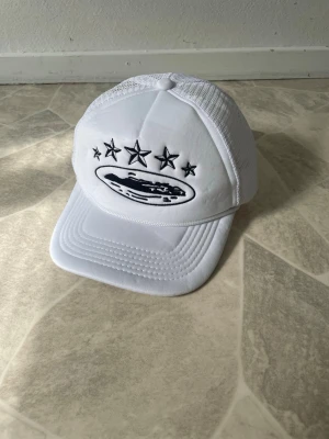 Vit meshkeps från Corteiz - Säljer en vit keps från Corteiz med svart broderad logga och stjärnor framtill. Kepsen har meshpaneler bak för extra ventilation och justerbar snapback. Perfekt för dig som gillar streetwear och vill ha en clean look.