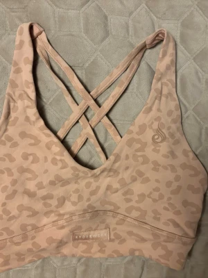 Beige leopardmönstrad sport-bh Ryderwear - Säljer en beige sport-bh från Ryderwear med diskret leopardmönster. Bh:n har korsade axelband i ryggen och en djup v-ringning framtill. Tillverkad i ett stretchigt och mjukt material som ger bra stöd och komfort.