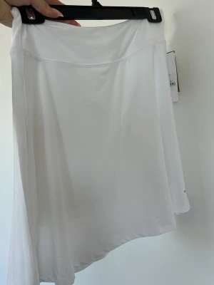 Vit kortkjol från Röhnisch  - Fräsch vit kortkjol från Röhnisch i ett sportigt och stretchigt material. Kjolen har en enkel och stilren design med diskret logga nertill. Perfekt för dig som gillar en clean och aktiv look. Aldrig använts!