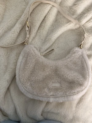 Vit fluffig axelremsväska från Zara - Supersöt axelremsväska från Zara i mjukt, fluffigt material och krämvit färg. Väskan har justerbar rem i konstläder och guldfärgade metalldetaljer. Perfekt för dig som gillar trendiga accessoarer med cozy vibe. Stängs med dragkedja 
