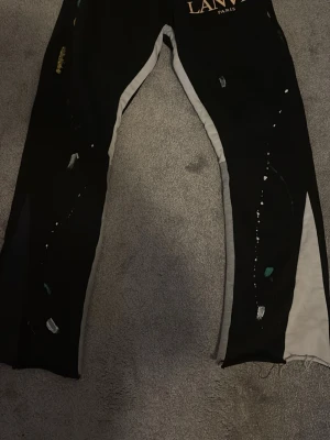 Svarta mjukisbyxor från Lanvin Paris - Säljer ett par svarta mjukisbyxor från Lanvin Paris med vita sidopaneler och färgstänk i grönt, vitt och gult. Byxorna har en avslappnad passform och tryckt logga framtill. Perfekta för en chill och trendig look.