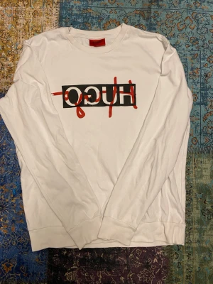 Vit långärmad tröja från Hugo Boss - Säljer en vit långärmad tröja från Hugo Boss med tryckt HUGO-logga i svart och rött framtill. Tröjan har rund halsringning och är i mjukt material, perfekt för en clean och modern look.