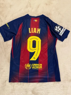 Barcelona matchtröja Nike blå/röd - Säljer en officiell FC Barcelona matchtröja från Nike i blått och rött med gula detaljer. Tröjan har trycket 'LIAM' och nummer 9 på ryggen, Champions League-märke på ärmen och Spotify-logga på bröstet. Tillverkad i lätt och ventilerande material.