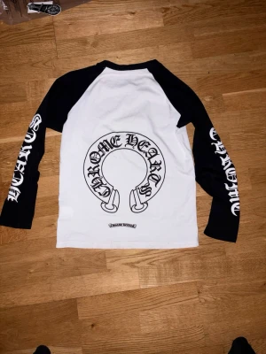 Chrome Hearts långärmad topp - Säljer en långärmad topp från Chrome Hearts med svartvita kontraster. Stora logotexter på båda ärmarna och ett stort tryck på ryggen. Klassisk raglanmodell med svart krage och ärmar, samt vit kropp. Perfekt för dig som gillar streetwear och statement-plagg.