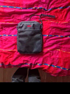 Svart axelväska från Levi's - Snygg svart axelväska från Levi's med justerbar axelrem och dragkedja. Väskan har en meshficka framtill och en diskret röd Levi's-logga. Tillverkad i slitstarkt tyg, perfekt för att bära det viktigaste. Enkel, stilren och passar till det mesta.