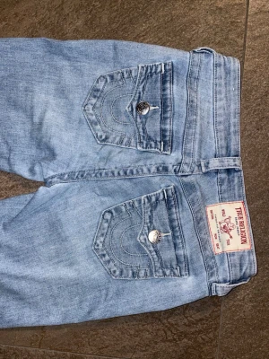 Ljusblå bootcut jeans från True Religion Becca - Säljer ett par ljusblå bootcut jeans från True Religion med klassiska kontrastsömmar och snygga fickdetaljer bak. Jeansen har en lätt tvättad look och är tillverkade i denim med stretch för skön passform. Perfekta för dig som gillar retrovibbar och ikonisk stil. 