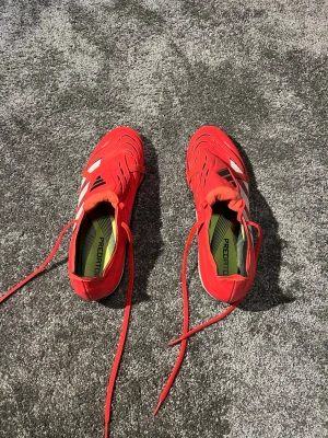 Adidas Predator röda fotbollsskor - Säljer ett par röda Adidas Predator fotbollsskor med svarta och vita detaljer. Skorna har snörning och en modern, sportig design med mönstrade linjer på ovandelen. Perfekta för dig som vill sticka ut på planen och få bra grepp om bollen.