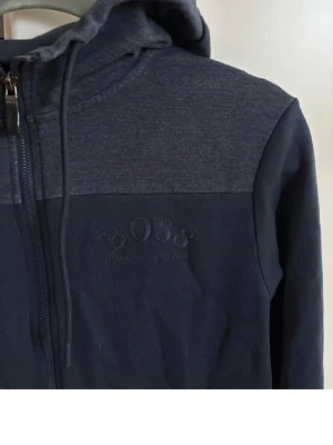 Mörkblå hoodie från Hugo Boss - Mörkblå hoodie från Hugo Boss med dragkedja framtill och huva med dragsko. Diskret broderad logga på bröstet och två fickor. Perfekt för en chill och stilren look.