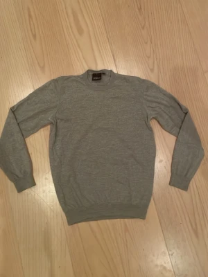 Grå stickad tröja Oscar Jacobson - Snygg grå/lite beige stickad tröja från Oscar Jacobson, tillverkad i Italien. Gjord av 100% merinoull och är enkel att matcha med andra kläder. Hör av er vid frågor eller funderingar