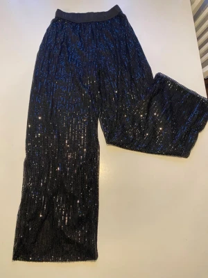 Svarta paljettmjukisbyxor med resår - Säljer ett par svarta mjukisbyxor täckta med glittrande paljetter. Byxorna har bred resår i midjan och dragsko för skön passform. Vida ben och mjukt tyg på insidan gör dem både snygga och bekväma. Perfekta för dig som vill sticka ut lite extra.