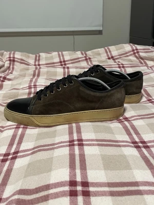 Lanvin skor  - Snygga sneakers i brunt mocka med svart tåparti i blankt material och beige sula. Klassisk låg modell med svarta skosnören och diskret design. Perfekta för dig som gillar stilrena och tidlösa skor. Skorna är i helt okej skick 6,5 av 10. Innersulan är trasig men kan nog fås med nya. Skriv vid minsta lilla