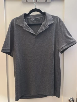 Mörkgrå piké från asos  - Klassisk mörkgrå pikétröja från asos med korta ärmar och mjuk krage. Tillverkad i bomullsmix för en bekväm känsla och enkel stil. Perfekt till jeans eller shorts för en clean look.