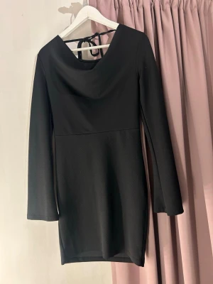 Svart långärmad kortklänning - Stilren svart kortklänning med långa ärmar och snygg snörning i nacken. Klänningen har en mjuk, något stretchig passform och en lätt draperad urringning. Perfekt för dig som gillar minimalistisk och trendig stil. Själva papperslappen är borta men helt ny, passar även S