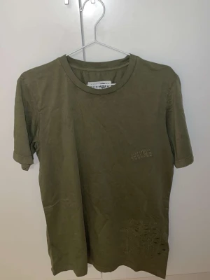 Margiela Olivgrön t-shirt  - Säljer en stilren olivgrön t-shirt från Maison Margiela med diskret broderad logga på bröstet. T-shirten har rund halsringning och korta ärmar. Tillverkad i mjuk bomull för en skön känsla och enkel att matcha med olika outfits.