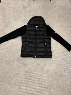 Svart cardiganjacka från Moncler - Svart cardiganjacka från Moncler med quiltad, glansig front och huva. Ärmarna är stickade i svart material och har Monclers klassiska logotyp på vänster ärm. Jackan har dragkedja framtill och två fickor med dragkedja. Skicka gärna frågor eller prisförslag ☺️