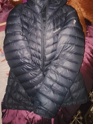 Svart dunjacka Peak Performance M - Snygg svart dunjacka från Peak Performance med huva och quiltad design. Jackan har dragkedja framtill och diskret logga på bröstet. Perfekt för kalla dagar och riktigt skön att bära tack vare det lätta dunmaterialet.